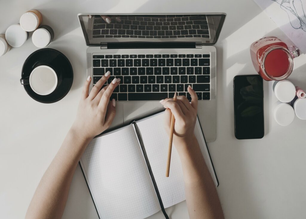 A person typing on a laptop while also holding a pencil to write ideas on a notebook.